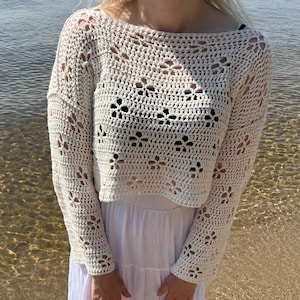 May include: Cream-colored, open-weave crochet top with long sleeves and a boat neck. The top features a floral pattern and is paired with a white dress. The background shows water and a sandy beach.
