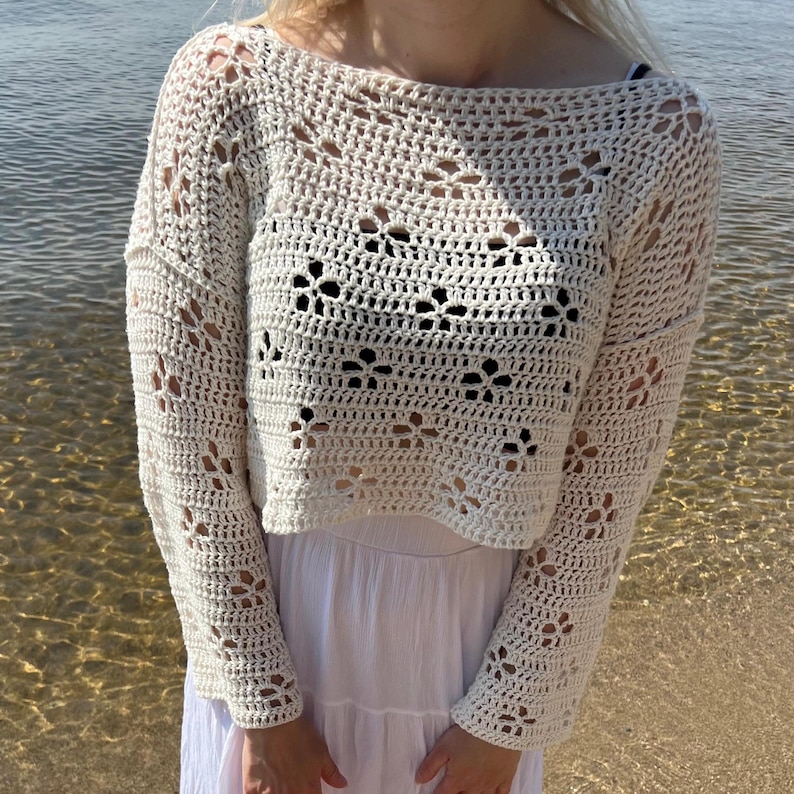 May include: Cream-colored, open-weave crochet top with long sleeves and a boat neck. The top features a floral pattern and is paired with a white dress. The background shows water and a sandy beach.