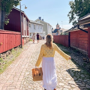 May include: A woman walking down a cobblestone street, wearing a yellow crochet top and a white maxi dress. She carries a brown paper bag. Buildings with red and light blue exteriors line the street, with trees and a clear blue sky in the background.