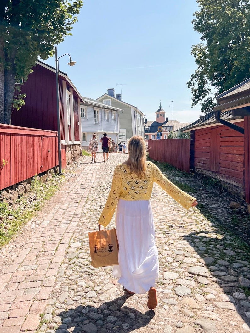 May include: A woman walking down a cobblestone street, wearing a yellow crochet top and a white maxi dress. She carries a brown paper bag. Buildings with red and light blue exteriors line the street, with trees and a clear blue sky in the background.
