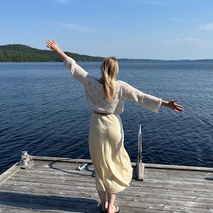 May include: A cream-colored, open-weave crochet top with long sleeves and a matching light yellow skirt. The outfit is worn on a wooden dock overlooking a lake. The person is wearing black sandals.