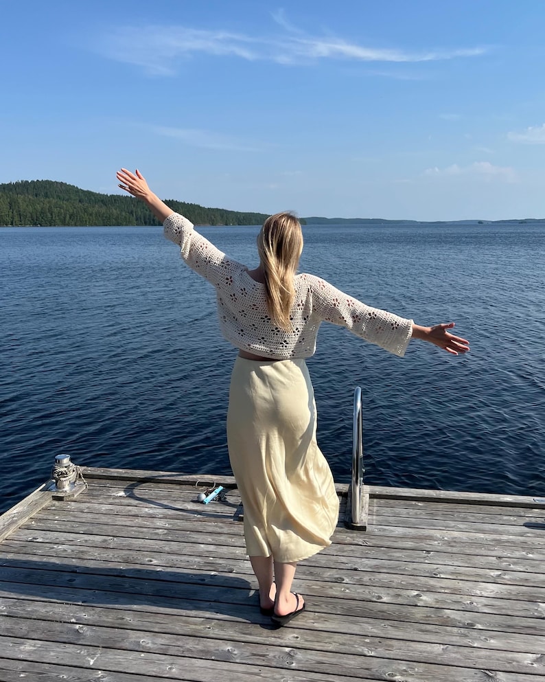 May include: A cream-colored, open-weave crochet top with long sleeves and a matching light yellow skirt. The outfit is worn on a wooden dock overlooking a lake. The person is wearing black sandals.