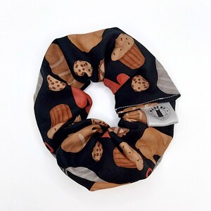 Może przedstawiać: Czarna scrunchie z brązowym i białym nadrukiem ciasteczek i ciastek. Scrunchie ma białą metkę z napisem "Made by Maurice".