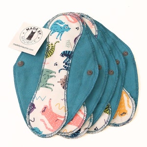 May include: A set of six teal reusable cloth pads with a white cotton top featuring a colorful cat print. The pads have a snap closure and the text "MADE BY MAURICE madebymaurice.com" is printed on a white tag.