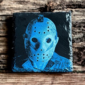 Op de afbeelding: Een blauwe en zwarte leisteen onderzetter met een afbeelding van een hockeymasker. Het masker heeft een witte omtrek en een zwarte achtergrond.
