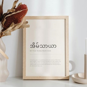 May include: A framed print with the Burmese word "အိမ်သာယာ" which translates to "Home Sweet Home" in English. The print is on a light beige background with a wooden frame. The print is sitting on a white surface with a candle holder and a vase of dried flowers.