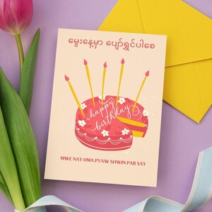以下が含まれることがあります： ピンクと白の誕生日カード。ピンクのケーキにろうそくが立てられており、「Happy Birthday」と「Mwe nay hma pyaw shwin par say」の文字が書かれています。