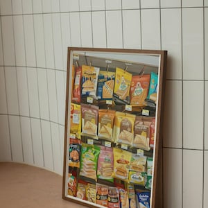 May include: A framed print featuring a display of packaged sandwiches. The sandwiches are in colorful packaging, with various brands and flavors visible. The print is in a brown wooden frame, set against a white tiled wall.