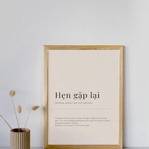 Può includere: Stampa incorniciata con la frase vietnamita "Hẹn gặp lại" in un design minimalista. Il testo nero è su sfondo crema, racchiuso in una cornice di legno chiaro. Un piccolo vaso con fiori secchi è in primo piano.
