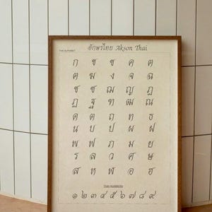 Pode incluir: Uma impressão emoldurada com o alfabeto e os números tailandeses. O texto "Thai Alphabet" e "Thai Numbers" é visível. A impressão está em uma moldura de madeira marrom, com texto preto sobre fundo creme.