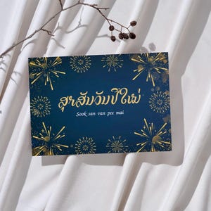 Puede incluir: Una tarjeta de felicitación azul marino con un diseño de fuegos artificiales dorados. La tarjeta tiene texto en un idioma extranjero y la traducción al inglés "Sook san van pee mai". Una ramita de una planta está en la esquina superior izquierda.
