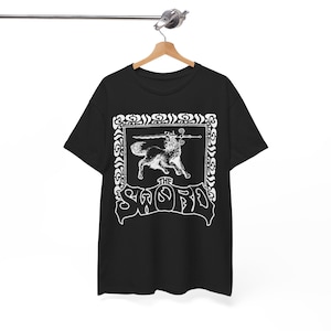 May include: Black t-shirt featuring a white graphic of a wolf holding a sword, framed by an ornate border. The word "THE SWORD" is displayed in a stylized font below the wolf image. The shirt is hanging on a wooden hanger.