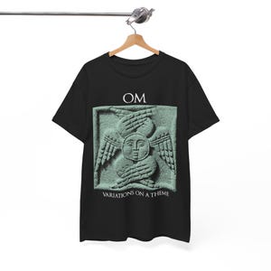 Puede incluir: Camiseta negra con un diseño gráfico verde claro de una cara con alas y manos, con el texto "OM" arriba y "VARIATIONS ON A THEME" abajo. La camiseta está colgada en una percha de madera.