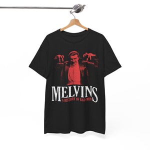 May include: Black t-shirt featuring a red graphic of a man with outstretched hands. The text "MELVINS" is displayed in white, with "A HISTORY OF BAD MEN" below it. The shirt is hanging on a wooden hanger.