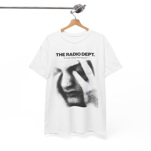 May include: White t-shirt with the text "THE RADIO DEPT." above a black and white image of a face with hands covering it. The text "Strange Things Will Happen" is below the band name. The shirt is hanging on a wooden hanger.