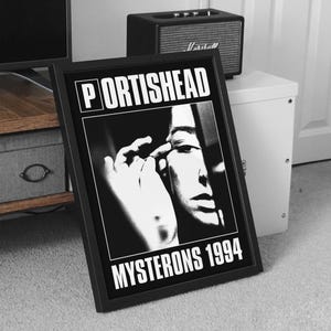 Puede incluir: Póster enmarcado en blanco y negro con el nombre de la banda "Portishead" y el texto "Mysteron 1994". La imagen del póster es un primer plano en escala de grises de un rostro y una mano. Un pequeño altavoz negro se encuentra detrás del póster.