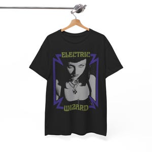 May include: Black t-shirt featuring a grayscale image of a woman with the text "ELECTRIC WIZARD" in green and purple. The design is framed by a purple lightning bolt shape. The shirt is hanging on a wooden hanger.