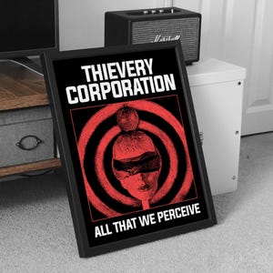 May include: Framed black and red art print with the text "THIEVERY CORPORATION" and "ALL THAT WE PERCEIVE". The artwork features a blindfolded face with a red target design in the background.