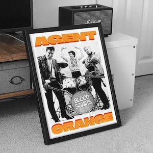 May include: A black and white photo of a band called "Agent Orange" with the words "Blood Stains" on a drum. The band members are wearing black clothing and the text is in orange.