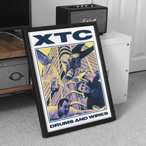 May include: Framed poster featuring the band XTC with the text "XTC" in navy blue and "DRUMS AND WIRES" in white. The image has a retro style with a yellow and purple color scheme. The poster is in a black frame.