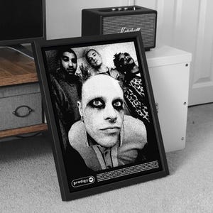 May include: Black and white framed poster featuring a group of people. The central figure has dark eye makeup and a nose ring. The poster is set against a neutral background with a speaker and cabinet visible.