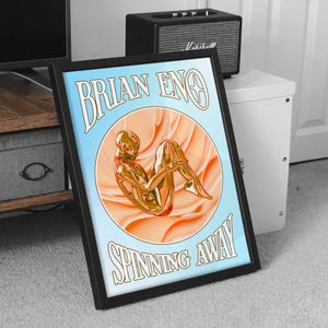 Può includere: Stampa artistica incorniciata con il testo "BRIAN ENO" e "SPINNING AWAY". L'opera raffigura una figura dorata in un design circolare su uno sfondo azzurro e pesca. La cornice è nera.