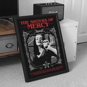 May include: A black and white framed print with a gothic design featuring a woman holding a skull. The text "THE SISTERS OF MERCY" is in red at the top of the print. The text "IN THE TEMPLE OF LOVE YOU HIDE TOGETHER BELIEVING PAIN AND FEAR OUTSIDE" is in red at the bottom of the print.