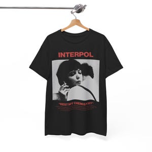 Peut inclure: T-shirt noir avec le nom du groupe "INTERPOL" en rouge au-dessus d'une image en noir et blanc d'une personne fumant. Le texte "REST MY CHEMISTRY" est imprimé en dessous de l'image.