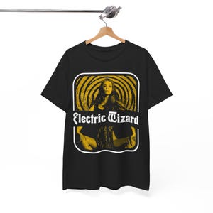 May include: Black t-shirt featuring a yellow and white graphic. The graphic includes a woman's image and the text "Electric Wizard" in white. The design is set within a rectangular frame with a white border. The shirt is hanging on a wooden hanger.