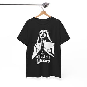 May include: Black t-shirt featuring a white graphic of a nun with clasped hands and the text "Electric Wizard" in a gothic font. The design is centered on the shirt, which is hanging on a wooden hanger.