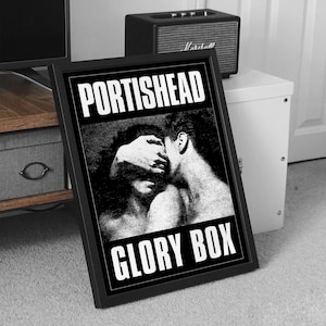 Puede incluir: Póster enmarcado en blanco y negro con el nombre de la banda "PORTISHEAD" y el título de la canción "GLORY BOX". La obra de arte representa dos figuras en un abrazo cercano. Un pequeño altavoz negro está sobre un armario blanco.