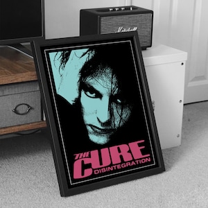 Könnte beinhalten: Gerahmtes Poster mit einem stilisierten Porträt einer Person mit schwarzen Haaren und einem türkisfarbenen Hintergrund. Der Text "THE CURE DISINTEGRATION" ist in Rosa gehalten. Das Poster befindet sich in einem schwarzen Rahmen.