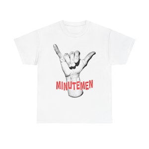 May include: White t-shirt featuring a grayscale illustration of a hand making the shaka sign, with the word "MINUTEMEN" in red block letters underneath. The shirt is a classic crew neck style.