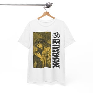 May include: White t-shirt featuring a religious image of Jesus Christ with a crown of thorns, carrying a cross. The text "GETHSEMANE" is printed vertically in black, along with a symbol. The shirt is hanging on a wooden hanger.