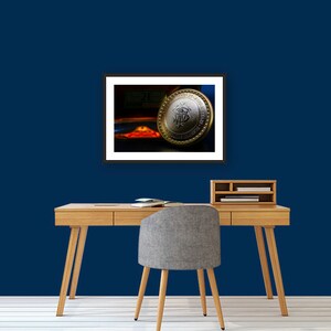 May include: A framed photograph of a silver and gold coin with a design on it. The coin is resting on a red and orange surface. The background is dark blue.