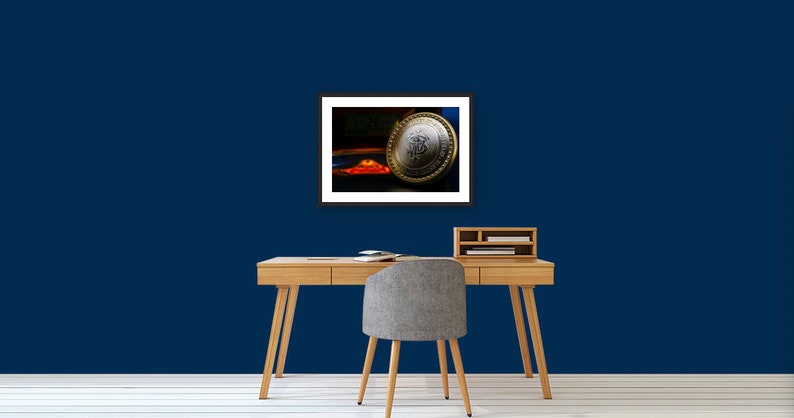 May include: A framed photograph of a silver and gold coin with a design on it. The coin is resting on a red and orange surface. The background is dark blue.