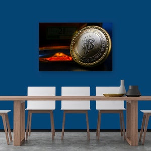 May include: A gold and silver coin with a design featuring a stylized letter "M" inside a circle. The coin is on a table with a blue wall in the background.