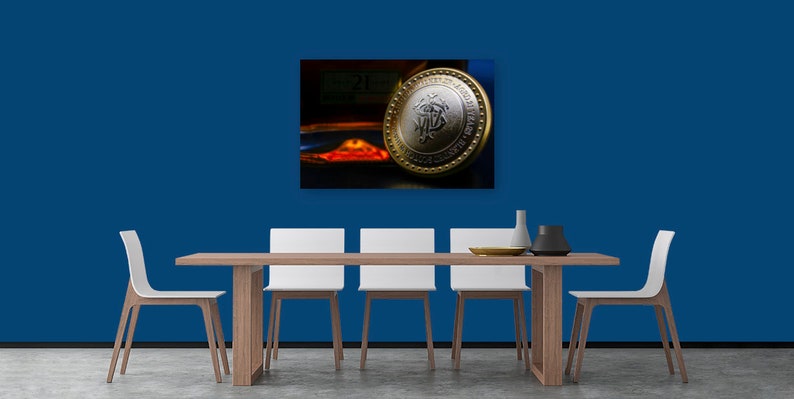 May include: A gold and silver coin with a design featuring a stylized letter "M" inside a circle. The coin is on a table with a blue wall in the background.