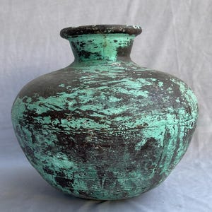 May include: A round vase with a wide body and a narrow neck, showing signs of age. The vase has a distressed finish, with a turquoise-green hue over a darker base. The rim is dark, and the overall effect is rustic and aged, with a vintage feel.