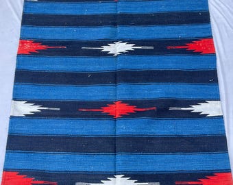 Puede incluir: Alfombra rectangular tejida con un patrón de rayas en tonos azules, negros, rojos y blancos. La alfombra tiene flecos a lo largo del borde inferior. El diseño presenta rayas horizontales con acentos geométricos.
