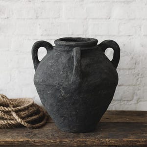 May include: A large, dark charcoal-colored vase with two handles. The vase has a textured surface and sits on a rustic wooden table. A coil of rope is visible to the left of the vase. The background is a white brick wall.
