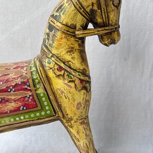 May include: A hand-carved wooden horse figurine, painted in shades of yellow and brown with decorative accents. The horse features a detailed saddle and bridle, with red, green, and white patterns. The figurine is a decorative item.