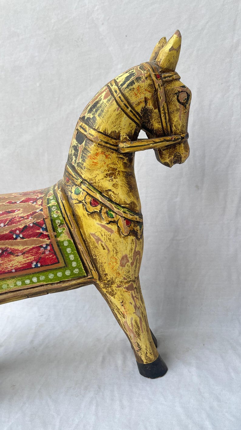 May include: A hand-carved wooden horse figurine, painted in shades of yellow and brown with decorative accents. The horse features a detailed saddle and bridle, with red, green, and white patterns. The figurine is a decorative item.