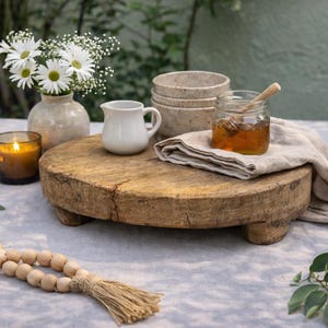 May include: A rustic wooden serving board holds a small white pitcher, a stack of beige bowls, and a jar of honey with a wooden dipper. A vase of white flowers and a lit candle are nearby. A beaded garland with a tassel is in the foreground.