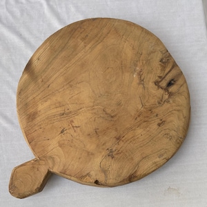 以下が含まれることがあります： 丸い木製のまな板。明るい茶色の木材で作られており、木目がはっきりと見えます。長方形の取っ手が側面から出ています。直径約30cmのこのまな板は、食品の調理に使用される可能性があります。