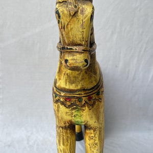 May include: A hand-carved wooden horse figurine, painted in a distressed yellow with black hooves. The horse has detailed features, including a bridle and decorative accents in red, green, and gold. A unique home decor item.