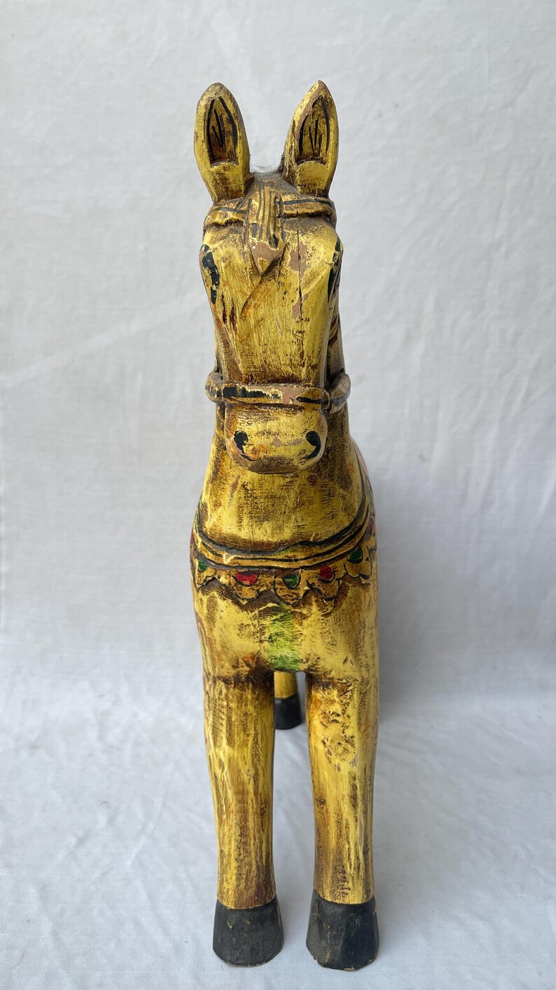 May include: A hand-carved wooden horse figurine, painted in a distressed yellow with black hooves. The horse has detailed features, including a bridle and decorative accents in red, green, and gold. A unique home decor item.