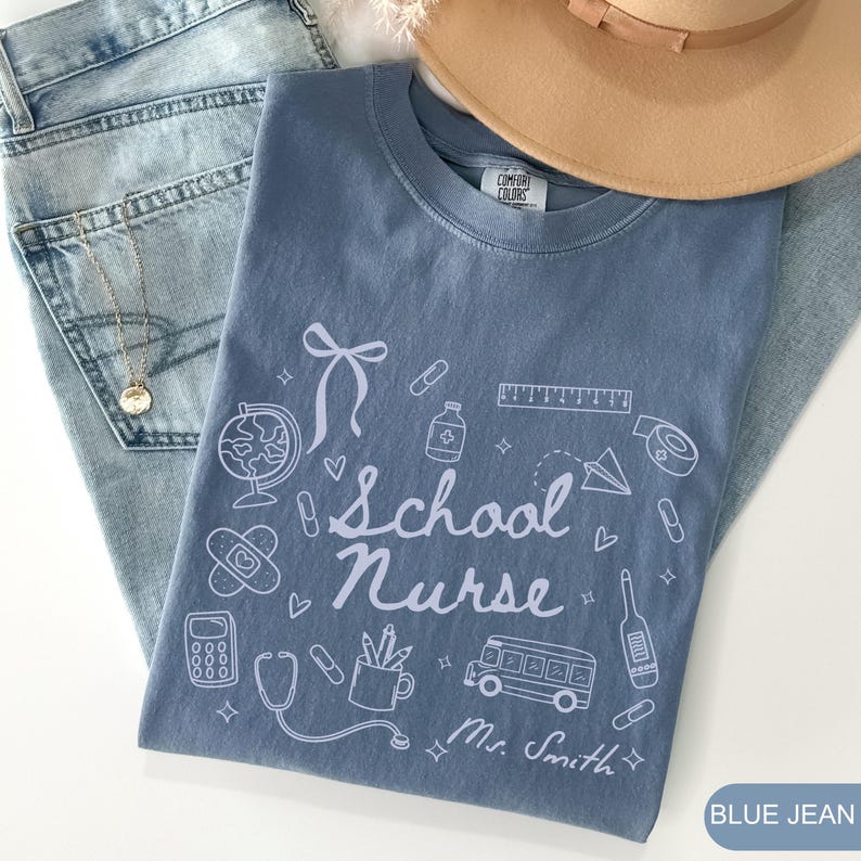 May include: A blue jean Comfort Colors t-shirt with the words "School Nurse" and related graphics. The shirt is paired with blue jeans, a gold necklace, and a tan hat. The name "Mr. Smith" is printed on the shirt.