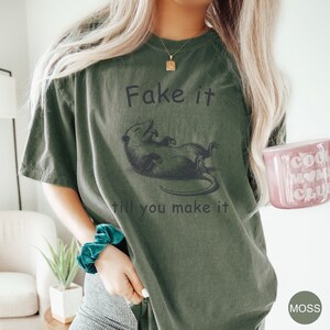 May include: A green t-shirt with a black graphic of a possum and the text "Fake it till you make it".