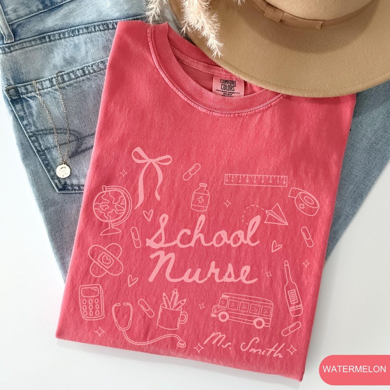 May include: A watermelon-colored t-shirt with a white graphic design. The design includes the words "School Nurse" and various school and medical-related illustrations. A hat and jeans are in the background.